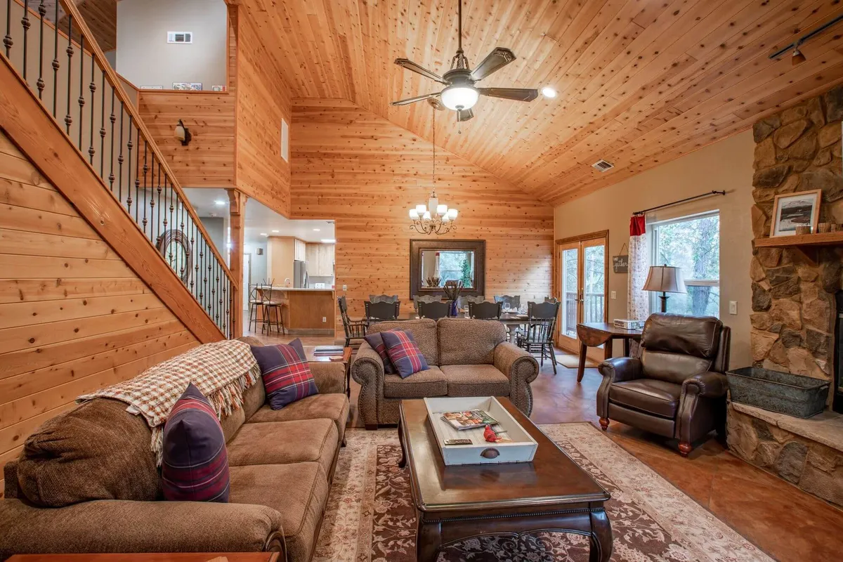 A spacious, open-concept living and dining area with a warm wooden ceiling. There’s a prominent stone fireplace beside a cozy, comfortable sofa, with a dining area visible in the background. The room is bathed in soft, inviting light, enhancing the welcoming ambiance.