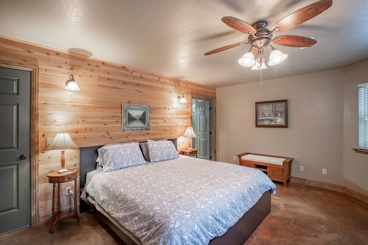 This image shows a cozy bedroom featuring a neatly made bed with decorative bedding, warm wood paneling on the walls, a ceiling fan, nightstands with lamps, and tasteful artwork. Sunlight filters in, creating a calm and serene atmosphere ideal for rest.