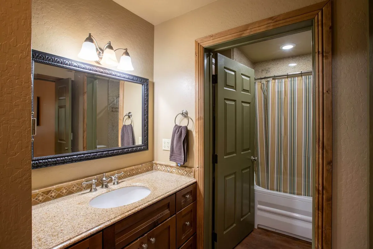 This bathroom features a sleek granite countertop, a wide mirror, wooden cabinetry, and a visible shower area. The space is styled for both functionality and comfort, with a cozy atmosphere suited for relaxation and daily routines.
