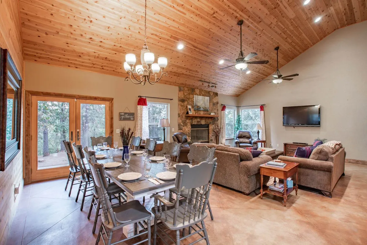 The image showcases a warm, inviting living and dining area with a rustic dining table set for gatherings, a glowing fireplace, plush sofa set, grand chandelier, and expansive windows letting in natural light and views of the surrounding forest.